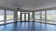 This single-floor space is defined by its striking curved exterior wall, lined with large windows fitted with roller shades that allow for flexible light control throughout the day. French doors with transom windows above occupy the center of this curved wall, opening directly onto a balcony with a metal railing and commanding views of surrounding greenery. The abundance of glazing ensures the interior is bathed in natural light, with treetop scenery visible from multiple angles, creating a seamless connection between the indoors and the outdoors.

The flooring throughout is a rich, dark hardwood, lending warmth and contrast to the neutral gray walls and white trim. The smooth, light-toned ceiling is equipped with a ceiling fan, providing both functionality and a subtle architectural detail. The open layout offers a generous, flexible living area without interior partitions, allowing the architectural features — particularly the panoramic window arrangement and the curved wall — to remain the focal points. The overall ambiance is airy, modern, and light-filled, with clean lines and a refined, understated aesthetic.