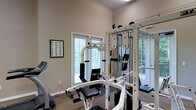 This single-floor fitness space is designed to support a comprehensive workout experience. At its centerpiece stands a Paramount multi-station cable machine, a robust, white-framed unit offering a variety of strength training options including cable pulls, a lat pulldown station, and adjustable pad attachments. Additional exercise equipment is visible throughout the room, including what appears to be a treadmill along the far wall, maximizing the available floor space.

The room features neutral beige walls paired with carpeted flooring, creating a clean and functional environment. Vaulted ceilings add a sense of openness to the space, preventing it from feeling confined despite the density of equipment. Natural light filters in generously through multiple windows fitted with horizontal blinds, providing views of surrounding greenery and contributing to an airy, welcoming atmosphere.

A framed instructional chart is mounted on the wall, offering exercise guides or muscle-group references for users. The overall layout is organized and practical, with electrical outlets conveniently placed along the baseboards. The space strikes a balance between functionality and comfort, making it suitable for regular fitness use.