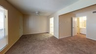 This single-floor unit presents a practical and straightforward layout designed for comfortable everyday living. The living area is generously sized, featuring wall-to-wall carpeting in a warm neutral tone that extends throughout the space. A ceiling fan with a light fixture is centrally positioned overhead, complementing the textured popcorn ceiling. A window along one wall allows natural light to filter in, offering a view of surrounding greenery. A closet and an additional door provide convenient access and storage directly from the living space.

The galley-style kitchen runs along two parallel walls and is finished with light tile flooring. White cabinetry with gold-toned hardware lines both sides, offering ample storage above and below the counters. White appliances, including a coil-burner range, refrigerator, and a sink with a single basin, are neatly integrated into the layout. A ceiling-mounted light fixture and a ventilation register keep the kitchen functional and well-lit.

The bathroom area features a vanity with a white countertop and cabinetry below, a towel bar, and access to a separate wet area housing a full bathtub with a showerhead. Tile flooring transitions into carpet within the vanity corridor, keeping the space clean and organized.
