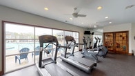 This fitness center is a comprehensive, single-floor facility designed to support a full range of workout routines. The space is bright and open, featuring large windows that flood the interior with natural light and offer views of an outdoor pool area, creating an energizing atmosphere for exercise. Rubber flooring throughout provides durability and comfort underfoot, while ceiling fans help maintain air circulation across the workout areas. The cardio section is well-appointed with multiple Life Fitness treadmills and elliptical trainers arranged to face the pool-facing windows, along with wall-mounted televisions for entertainment during workouts. A large wall mirror expands the visual depth of the space and allows users to monitor their form. The strength training area includes a cable machine, a multi-function weight machine, and additional resistance equipment, providing options for a variety of targeted exercises. A dedicated free weights section features a full rack of dumbbells alongside flat benches, catering to both beginner and advanced lifters. Elegant wood-framed glass French doors connect to an adjacent room, which appears to house additional equipment and storage. Recessed lighting complements the natural light, ensuring the space remains well-lit at all times. Overall, the fitness center delivers a clean, functional, and motivating environment.