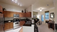 This single-floor, one-bedroom, one-bathroom unit features an open-concept design that maximizes its well-proportioned square footage. The kitchen is a standout element, outfitted with warm wood cabinetry extending to the upper walls, dark granite countertops, and stainless steel appliances including a range, built-in microwave, and double-basin sink. A breakfast bar with seating provides a casual dining option while maintaining visual connection to the living area. Track lighting over the kitchen ensures ample task illumination.

The living area flows naturally from the kitchen, featuring ceiling-height walls with a neutral color palette accented by a sage green feature wall. A ceiling fan with integrated lighting provides both comfort and ambient light. Large windows allow natural light to filter into the space, creating a bright and welcoming atmosphere. The carpeted living area offers a comfortable contrast to the kitchen's hard flooring. Crown molding details add a refined architectural touch throughout, contributing to the overall polished and functional character of the unit.