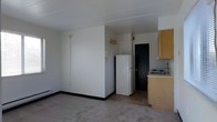 Located at 300 W 3rd Street in Bridgeport, PA, this single-floor unit offers a straightforward and functional layout designed for efficient everyday living. The open living area features neutral-toned carpeting that creates a clean, understated foundation, while white walls and a flat ceiling keep the space feeling bright and simple. A baseboard heating unit runs along the lower wall, ensuring year-round comfort. The kitchen area is positioned along one side of the unit and includes light wood cabinetry providing ample storage, a countertop with an integrated sink, and a full-size refrigerator that sits adjacent to the transition between the carpeted living space and tiled kitchen floor. The tile flooring in the kitchen zone offers a practical, easy-to-clean surface. A wall-mounted light fixture with a gold-toned finish adds a decorative touch near the kitchen. A dark interior door leads to an additional room or utility space beyond the main living area, adding a degree of privacy and separation within the unit. Overall, the space presents a no-frills, utilitarian design that prioritizes functionality and practicality, making it a straightforward option for those seeking a simple, well-located residence in Bridgeport, PA.