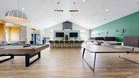 Located at 310 Northbend Drive in Charlotte, NC, this single-story amenity space is a thoughtfully designed community hub offering a diverse range of functional zones. The interior is characterized by soaring vaulted ceilings, light wood-look tile flooring, and a vibrant color palette of greens, teals, golds, and whites that energize the space throughout.

The central gathering area serves as the social heart of the space, anchored by a large U-shaped bar counter lined with contemporary upholstered barstools. Twin flat-screen TVs are mounted against a soft teal accent wall, and a coffee station and vending machines provide convenient refreshments. Dramatic pendant chandeliers with exposed Edison bulbs hang from the high ceiling, adding warmth and visual interest.

Adjacent to the lounge is a recreation area featuring a full-size pool table beneath a dome pendant light, alongside intimate booth-style seating with semi-private dividers — ideal for casual gatherings or focused work. A bright, welcoming corridor with bold green accent walls, abstract artwork, and yellow lounge seating connects the main zones seamlessly.

A dedicated conference room offers a more private setting, furnished with a long white table, ergonomic seating, floor-to-ceiling windows, and a wall-mounted display screen. A business services nook with printing capabilities and a private office space round out the functional offerings, making this a well-equipped and inviting community facility.