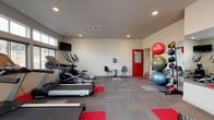 Located at 2555 Bella Colla Lane in Richland, WA, this single-floor fitness center is designed to support a well-rounded workout routine in a bright, welcoming environment. The space features a modern aesthetic with neutral-toned walls, recessed lighting, and a patterned carpet floor accented with bold red tiles that add energy to the room.

The cardio section is generously equipped with multiple treadmills, elliptical trainers, and a stationary recumbent bike, all positioned along a wall of large windows fitted with horizontal blinds. These windows provide ample natural light and outdoor views, creating an open and airy atmosphere during workouts. Wall-mounted flat-screen televisions are positioned throughout the space for entertainment.

A multi-station cable and weight machine anchors the strength training area, complemented by a full-length wall mirror that enhances the sense of space. A rack of exercise balls in various sizes adds versatility for core and flexibility training, while red workout mats on the floor provide designated zones for stretching and floor exercises.

The space also includes a small seating area with modern chairs, a hand sanitizer station, and a waste bin, reflecting attention to cleanliness and user comfort. A bold red interior door with frosted glass panels leads to an adjacent room, adding a distinctive design accent. Two exterior glass doors provide easy access from outside.
