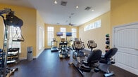 Located at 2067 Springdale Ln in Gallatin, Tennessee, this well-appointed fitness center is part of the Revere Luxury Living community. The single-floor facility is thoughtfully designed to support a comprehensive workout experience in a bright, welcoming environment.

The space is bathed in natural light, courtesy of numerous windows fitted with white blinds that line the warm yellow walls. Recessed lighting and a ceiling fan overhead ensure the room remains comfortable and well-lit at all hours. The durable speckled rubber flooring throughout provides both safety and cushioning during exercise.

Cardio equipment is a highlight, with multiple True-brand treadmills, recumbent bikes, upright stationary bikes, and elliptical-style machines arranged across the room. Wall-mounted flat-screen televisions are positioned for easy viewing from the cardio stations, adding an entertainment element to workouts.

A dedicated free weights area features a rack of dumbbells in varying sizes alongside an adjustable workout bench. A large wall mirror expands the sense of space and allows for proper form checks. A multi-station cable and strength machine rounds out the resistance training options.

The facility is accessible via a glass-paneled exterior door and includes clearly marked emergency exits, reflecting a focus on safety. The overall ambiance is clean, organized, and inviting, making this fitness center a functional and attractive amenity for residents.