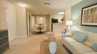 This single-floor residence offers an open-concept floor plan that seamlessly integrates the living, dining, and kitchen areas into one cohesive and functional space. The living room is anchored by a light-toned sofa accented with decorative pillows and paired with a wooden coffee table, creating a warm and inviting atmosphere. A soft sage green accent wall adds a subtle pop of color, complementing the overall neutral palette of beige and cream tones found throughout. The dining area features a round table surrounded by upholstered chairs beneath a stylish pendant light, with a framed artwork and a warm accent wall serving as an elegant backdrop. The kitchen is positioned adjacent to the dining space and is accessible via a half-wall breakfast bar with a granite-style countertop, providing both separation and connectivity. White cabinetry and under-cabinet lighting contribute to a clean, bright kitchen aesthetic. Consistent light-toned carpeting flows throughout the main living areas, enhancing the sense of continuity and space. Crown molding and column details add refined architectural touches, while ample ceiling-mounted lighting ensures the entire space remains well-lit. A hallway visible from the living area suggests additional private rooms beyond the main open-plan zone.