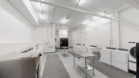 Located at 250 W 5th St in downtown Cincinnati, Ohio, this shared laundry facility is designed with functionality and efficiency in mind. The single-floor space features a clean, bright interior finished in crisp white throughout, with exposed overhead piping and fluorescent lighting that ensures the entire room is well-illuminated.

The facility is equipped with multiple commercial-grade washers and dryers — including both top-load and front-load machines — arranged along the walls to maximize usable floor space. Stainless steel folding tables are positioned centrally, providing ample surface area for sorting and folding laundry. Gray area rugs define the primary work zones, adding a modest layer of comfort underfoot on the polished concrete floors.

A row of black storage lockers is situated at the rear of the space, complemented by a seating area with a metal bench for waiting. Vending and digital payment kiosks are conveniently accessible, supporting a modern, cashless laundry experience. Wall-mounted utility panels and a ceiling fan further contribute to the room's practical layout.

Double doors with frosted glass panels provide access to and from the facility, enhancing the sense of openness. The overall ambiance is utilitarian yet orderly, reflecting a well-maintained communal space designed to serve the needs of a multi-unit residential building efficiently.