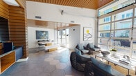 Located at 45 E Reed St in San Jose, California, this single-floor communal lounge space showcases a thoughtfully designed modern interior that seamlessly blends warmth and contemporary style. The standout architectural feature is the striking slatted wood ceiling, which adds warmth and texture while complementing the crisp white walls throughout. Underfoot, large-format hexagonal tiles in varying shades of gray and cream create a bold, geometric pattern that anchors the entire space.

Natural light is a defining characteristic here, pouring in through full-height glass walls and an oversized glass garage-style door that opens the interior to an outdoor terrace and courtyard beyond. The transparency of these walls offers views of lush greenery and the surrounding urban landscape, reinforcing a strong indoor-outdoor connection.

The lounge is furnished with woven rattan lounge chairs accented with soft blue cushions, a contemporary gray sofa, and clean-lined white tables with warm wood bases — creating inviting conversation areas throughout. Curated wall art, including abstract and minimalist pieces, adds personality and a gallery-like quality to the space.

Architectural columns clad in stacked wood panels serve as design focal points, further tying together the natural material palette. The overall ambiance is bright, airy, and welcoming — ideal as a gathering and relaxation space.
