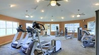 This fitness center is designed to support a full range of workout activities within a spacious, single-floor layout. The room features a gently vaulted ceiling fitted with multiple ceiling fans and recessed lighting, ensuring comfortable airflow and consistent illumination throughout the space. Warm terracotta-toned walls give the room an energizing yet welcoming atmosphere.

Natural light streams in through a series of windows positioned around the perimeter of the room, offering views of the surrounding greenery and creating an open, airy feel. The patterned carpet flooring adds a layer of comfort underfoot while defining the workout area.

The facility is outfitted with a variety of cardio equipment, including elliptical trainers and treadmills, positioned to take advantage of the natural light and open sightlines. Strength training stations are arranged along the windowed walls, providing a range of resistance and cable-based exercise options. The thoughtful layout allows for ample circulation space between machines, supporting safe and comfortable use by multiple visitors simultaneously.

Overall, the fitness center combines functional equipment placement with a well-designed interior environment, making it a practical and pleasant space for daily exercise routines.