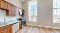 Located in Fall River, MA, this single-floor, one-bedroom, one-bathroom unit offers a well-designed layout with modern finishes throughout. The combined kitchen and living area serves as the heart of the unit, featuring warm-toned shaker-style cabinetry, granite countertops, and a full suite of stainless steel appliances including a refrigerator, range, microwave, and dishwasher. A track lighting fixture overhead adds a contemporary touch, while impressively tall ceilings lend a sense of openness and volume that extends throughout the space. Light-colored hardwood-style flooring flows seamlessly across all rooms, enhancing the bright, cohesive aesthetic. Oversized, multi-pane windows allow abundant natural light to fill both the kitchen/living area and the bedroom, which offers a comfortably sized layout and a peaceful, airy feel. The bathroom is well-appointed with granite countertops, wood-toned cabinetry, and the added convenience of a stacked in-unit washer and dryer. A hallway connects the rooms efficiently, with two closets providing practical storage solutions. Neutral wall tones and clean architectural lines give the unit a move-in-ready, contemporary character throughout.