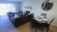 This single-floor residence seamlessly blends a living area and dining space into one open, functional layout. The living area is anchored by a classic white-mantled fireplace, which serves as a focal point and adds warmth to the room. Decorative framed artwork and a round mirror adorn the mantle, lending a curated, homey feel. A dark sofa paired with a patterned accent chair creates a comfortable seating arrangement, complemented by a modest side table and floor lamp that provide soft ambient lighting. Natural light filters in through a window, brightening the neutral-toned walls and carpet flooring that extend throughout the space. Adjacent to the living area, the dining space features a round dark-wood table surrounded by matching slatted-back chairs, set with white dinnerware for an inviting, ready-to-use presentation. A large decorative round mirror on the dining wall adds depth and visual interest. The open-plan design encourages easy flow between areas, making the space feel connected and versatile. The overall ambiance is warm, understated, and practical, suited for everyday living and casual entertaining.
