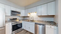 This single-floor residence offers a practical and well-designed layout with a clear separation between the kitchen and living areas. The kitchen is a standout feature, equipped with a full suite of stainless steel appliances including a range, over-the-range microwave, and dishwasher. White shaker-style cabinetry extends to the upper walls, providing generous storage, while granite-style countertops and under-cabinet lighting add a refined touch. Wood-look flooring runs throughout the kitchen, complementing its clean, modern aesthetic. A pass-through opening connects the kitchen to the living area, creating an open, communicative feel between the two spaces. The living area is generously sized, finished with neutral-toned carpet and light-colored walls that contribute to a bright, airy atmosphere. Windows fitted with vertical blinds allow natural light to filter in from the exterior. The bedrooms are carpeted and finished with the same neutral palette, offering comfortable, versatile spaces. One bedroom features a window with an outdoor view of surrounding greenery. The bathroom includes a marble-style vanity countertop with white cabinetry, wood-look flooring, and a separate water closet, providing a functional and organized layout throughout.