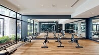 Located at 1650 NE 124th St in North Miami, FL, this Yoga & Spin Room is a thoughtfully designed fitness studio offering a versatile space for cycling, yoga, and Pilates. The studio features warm hardwood flooring throughout, creating a clean and energizing environment suited for a variety of workout disciplines. Large floor-to-ceiling glass windows and wall panels line the perimeter, flooding the space with abundant natural light and providing open views of lush outdoor greenery, bringing an inviting connection to the outdoors. The interior design follows a modern aesthetic with a striking contrast of dark navy accent walls and crisp white ceilings, accented by recessed lighting. Full-length wall mirrors expand the visual depth of the room while allowing practitioners to monitor their form. The studio is equipped with multiple stationary spin bikes arranged for group cycling sessions, as well as Pilates reformer equipment positioned near the window-lined walls. Glass partition walls provide visual connectivity to the adjacent main fitness area, where additional cardio and strength equipment is visible. Stability balls are neatly stored on dedicated racks near the entrance. The overall ambiance is sleek, modern, and motivating, making this studio a premium fitness destination within the broader facility.