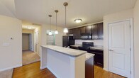 Located at 1300 Community Way in Lancaster, Ohio, this single-floor residence offers a well-designed layout with a cohesive blend of functional and comfortable living spaces. The kitchen is a standout feature, showcasing rich dark espresso cabinetry, light stone-look countertops, and black appliances including an electric range, microwave, and refrigerator. A peninsula with pendant lighting overhead provides additional prep space and serves as a natural divide between the kitchen and the adjoining living area. Hardwood-style flooring runs throughout the kitchen and entry zones, while plush carpeting extends into the living room and bedrooms. The living room impresses with vaulted ceilings that lend a sense of openness and height, complemented by natural light streaming through large windows. The bedrooms are generously sized, each featuring vaulted or raised ceilings and carpeted floors, contributing to a warm and quiet atmosphere. The unit includes two bathrooms: one full bath with a sliding glass-door shower stall and dark vanity cabinetry, and a second bathroom with a single-sink vanity and tile flooring. A dedicated laundry area with stacked washer and dryer hookups adds everyday convenience. The overall aesthetic is clean and neutral, with warm beige tones throughout.