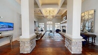 Located in Atlanta, GA, this single-floor space offers a sophisticated blend of classic architectural design and refined interior detailing. The grand living room commands immediate attention with its soaring coffered ceilings, an ornate orb chandelier, and rich hardwood floors that flow throughout. Stone-clad columns frame the entrance to the main seating area, where a floor-to-ceiling natural stone fireplace with a white wood mantel serves as a striking focal point. Arched French doors and large windows flood the space with natural light and offer views of the outdoors. Decorative wrought iron gates open to a spacious game room area, complete with a billiard table and additional seating nooks defined by arched doorways. The kitchen is equally impressive, featuring white cabinetry, granite countertops, stainless steel appliances, and a standout stone-faced island with bar seating. An ornately carved range hood and pendant crystal lighting add a touch of elegance. Throughout the space, soft blue-gray walls, stone accents, and carefully curated details create a warm, upscale ambiance ideal for both entertaining and everyday use.