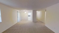 This single-floor space features an open-concept layout that creates a seamless flow between the living and dining areas. The main living space is finished with neutral beige carpeting throughout, contributing to a clean and understated aesthetic. A ceiling fan is centrally mounted, providing both ventilation and a functional design element.

A structural column divides the open floor plan, subtly defining separate zones within the shared space while maintaining an airy, connected feel. Sliding glass doors lined with vertical blinds allow natural light to filter into the room and likely provide access to an outdoor area.

The walls are painted in a light, neutral tone, enhancing the brightness of the interior. Visible from the main living area, the kitchen features white cabinetry and appears to connect directly to the open living space, reinforcing the overall open-plan design.

The textured ceiling adds a subtle architectural detail consistent throughout the space. Overall, the layout is practical and versatile, offering a blank canvas that can accommodate a variety of furniture arrangements and interior styles.
