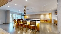 Located at 315 First Ave NE in Minneapolis, this generously sized single-floor amenity space offers a sophisticated blend of social, recreational, and lounge areas across an open, thoughtfully designed layout. The gourmet kitchen serves as a centerpiece, featuring a large island with bar seating, a striking stainless steel range hood, warm wood cabinetry, polished concrete floors, and a built-in banquette — ideal for gatherings and entertaining. A secondary kitchenette provides additional prep space with sleek white cabinetry, open wood shelving, and stainless steel appliances set against a textured tile backsplash. The lounge area is distinguished by a dramatic rope partition that defines the space without closing it off, complemented by floor-to-ceiling windows that flood the room with natural light and offer views of the surrounding neighborhood. A dedicated media lounge features a large-screen TV, plush reclining chairs, and a dark, moody ceiling that enhances the cinematic atmosphere. The game and social room includes a billiards table, high-top seating, exposed wood beam ceilings, and a linear fireplace with stone surround. A private dining area with wrap-around banquette seating and pendant lighting rounds out this versatile and well-appointed communal destination.