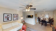 This single-story space in Denton offers a well-designed, open-concept layout that promotes a sense of flow and connectivity throughout. The living area serves as the central hub, featuring plush carpeted flooring, neutral-toned walls, and a ceiling fan with integrated lighting that provides both comfort and illumination. A half-wall partition elegantly separates the living space from the kitchen, which is visible in the background and features warm wood-toned cabinetry. The pass-through design of the partition allows for easy interaction between the kitchen and living areas while maintaining a defined sense of space. Adjacent to the living area, a dedicated dining space accommodates a dark-toned table and chairs, ideal for everyday meals or entertaining. Framed artwork on the walls adds a decorative touch, contributing to the overall warm and welcoming ambiance. The layout is practical and thoughtfully arranged, making efficient use of the available space while ensuring each area serves its purpose comfortably. The consistent flooring, cohesive color palette, and interconnected room design create a unified and inviting atmosphere throughout this single-floor residence in Denton.