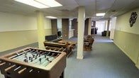 This single-floor communal space is designed for leisure and social gathering, offering an open and versatile layout ideal for group activities and relaxation. The room is anchored by several structural columns, some clad in brick, which add a distinct architectural character to the space. Overhead, a drop ceiling with recessed fluorescent lighting provides consistent, even illumination throughout.

The lounge area is furnished with a mix of wooden chairs and sofas arranged around coffee tables, creating comfortable conversation nooks. A foosball table takes center stage near the front of the room, making it a natural focal point for recreational activity. Toward the back, a counter or service area is visible, accompanied by what appears to be a lounge or seating alcove with vertical blinds allowing for natural light.

The floor is covered in a durable, neutral-toned carpet suitable for high-traffic use. The overall layout is open and unobstructed, allowing for flexible use of the space. With its combination of casual seating, game amenities, and a welcoming atmosphere, this communal area is well-suited for use as a resident lounge, student common room, or shared recreational facility.