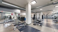 Located at 5720 Fishers Lane in Rockville, MD, this fitness center offers a generously sized, single-floor exercise space designed to support a full range of workout needs. The open layout is thoughtfully organized, with distinct zones for strength training and cardiovascular exercise, allowing for efficient use of the facility.

The strength training area is well-equipped with cable machines, multi-functional cable stations, a variety of free weights including a full dumbbell rack, adjustable benches, a punching bag, and a dedicated stretching and functional training zone featuring rubber flooring for safety and comfort. Large wall mirrors throughout amplify the sense of space and allow users to monitor their form.

The cardio section features a row of treadmills and step machines neatly arranged along a railed platform, providing an organized and accessible workout environment. Wall-mounted televisions are positioned throughout the facility for entertainment during workouts.

Architecturally, the space features exposed silver HVAC ductwork running along the ceiling, structural columns, and a mix of large-format tile and rubber flooring. Recessed and flush-panel ceiling lighting provides bright, even illumination throughout. Natural light enters through street-facing windows, and select accent walls display exposed brick and motivational graphics, contributing to an energetic and modern industrial ambiance.
