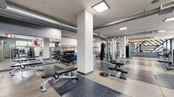 Located at 5720 Fishers Lane in Rockville, MD, this fitness center offers a generously sized, single-floor exercise space designed to support a full range of workout needs. The open layout is thoughtfully organized, with distinct zones for strength training and cardiovascular exercise, allowing for efficient use of the facility.

The strength training area is well-equipped with cable machines, multi-functional cable stations, a variety of free weights including a full dumbbell rack, adjustable benches, a punching bag, and a dedicated stretching and functional training zone featuring rubber flooring for safety and comfort. Large wall mirrors throughout amplify the sense of space and allow users to monitor their form.

The cardio section features a row of treadmills and step machines neatly arranged along a railed platform, providing an organized and accessible workout environment. Wall-mounted televisions are positioned throughout the facility for entertainment during workouts.

Architecturally, the space features exposed silver HVAC ductwork running along the ceiling, structural columns, and a mix of large-format tile and rubber flooring. Recessed and flush-panel ceiling lighting provides bright, even illumination throughout. Natural light enters through street-facing windows, and select accent walls display exposed brick and motivational graphics, contributing to an energetic and modern industrial ambiance.