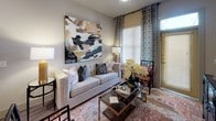 This thoughtfully designed single-floor residence blends contemporary style with comfortable living across a well-organized open layout. The kitchen is a standout feature, showcasing dark charcoal shaker-style cabinetry, gleaming quartz countertops, a metallic penny-tile backsplash, and a full suite of stainless steel appliances including a gas range, microwave, refrigerator, and dishwasher. A central island with pendant lighting provides additional prep space and casual seating, while wood-look flooring ties the kitchen seamlessly into the main living area. The living room is bright and welcoming, with large windows dressed in patterned drapes that allow natural light to fill the space. The bedroom offers plush carpeting, generous window coverage with blinds and curtains, and a sliding barn door detail that adds a modern farmhouse touch. A decorative headboard wall and ample space for furniture make it a restful retreat. The bathroom continues the refined aesthetic with gray cabinetry, quartz vanity surfaces, wood-look flooring, and a glass-enclosed walk-in shower. Neutral wall tones and curated décor details throughout lend the entire residence a cohesive, polished ambiance that is both functional and visually appealing.