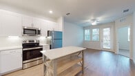 This well-appointed single-floor residence offers a clean, modern aesthetic throughout its thoughtfully designed layout. The open-concept kitchen serves as the heart of the home, outfitted with crisp white shaker-style cabinetry, a white subway tile backsplash, quartz countertops, and a full suite of stainless steel appliances including a refrigerator, range, microwave, and dishwasher. A freestanding kitchen island with a quartz top and lower shelf provides additional prep space and storage. Light wood-tone flooring extends through the kitchen and flows seamlessly into the adjoining living area, which features a ceiling fan and a glass door that invites natural light from outside. The bedrooms are finished with soft neutral carpet, offering a comfortable and quiet retreat. One bedroom includes dual windows with slatted blinds that frame views of the surrounding neighborhood, along with a ceiling fan overhead. The full bathroom is fitted with wood-look flooring, white cabinetry with stone countertops, a framed mirror, and a tiled tub-shower combination. A dedicated in-unit laundry room houses a full-size top-load washer and dryer pair with overhead wire shelving for added utility. The overall palette of whites, soft neutrals, and warm wood tones creates a bright and cohesive living environment.