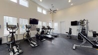 This single-floor fitness center is thoughtfully designed to accommodate a wide range of workout needs in a bright, open environment. The main exercise area boasts soaring double-height ceilings that create an airy, expansive atmosphere, enhanced by an abundance of natural light streaming through numerous windows fitted with blinds. Large ceiling fans help maintain a comfortable climate throughout the space.

The workout floor is finished with durable dark rubber flooring and houses a comprehensive selection of equipment, including treadmills, elliptical machines, a recumbent bike, a multi-station cable and weight machine, a rowing machine, and a full rack of dumbbells. Multiple wall-mounted flat-screen televisions are positioned throughout the room for entertainment during workouts. A large wall mirror in the strength training section expands the sense of space and allows for form monitoring.

Adjacent to the main gym floor is a dedicated children's room, thoughtfully separated by a glass partition wall that maintains visibility between spaces. This room features soft, colorful modular carpet tiles, a wall-mounted television, and an interactive activity panel, making it a welcoming space for young visitors. A door from the fitness area provides direct access to an outdoor area, further connecting the facility to its surroundings.
