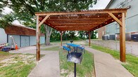 This outdoor communal space centers around a generously sized wooden pergola constructed with stained timber beams and a slatted lattice roof strung with café-style globe lights, creating an inviting atmosphere for daytime and evening gatherings. Beneath the pergola, a blue picnic table provides seating for groups, while pedestal-mounted charcoal grills are conveniently positioned for outdoor cooking and entertaining.

Concrete pathways extend outward from the pergola, connecting the various areas of the grounds. The surrounding landscape features a mix of open grassy areas and sandy patches, bordered by a low concrete retaining wall that defines the property boundary. A large, mature shade tree anchors the space near the pergola, contributing natural character and canopy coverage.

Adjacent to the main gathering area, additional structures are visible — including low-profile brick buildings with sage-green metal siding accents and cedar-shingled roof lines — alongside utility and storage outbuildings. Open ground extends toward a dense treeline at the rear of the property, offering a natural green backdrop. The overall setting is practical and functional, designed to support outdoor recreation, social gatherings, and casual use in a low-maintenance, open-air environment.