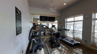 This single-story fitness center provides a well-equipped space for cardiovascular workouts. The room features an array of cardio machines, including treadmills, an elliptical, and a stationary recumbent bike, arranged to maximize the available floor space. A large wall-mounted mirror with a wood-trimmed rail spans one side of the room, visually expanding the space while allowing users to monitor their form during exercise. Two flat-screen televisions are mounted above the mirror, offering entertainment options during workouts. The ceiling hosts a white fan that provides air circulation throughout the room. Natural light filters in through a window fitted with horizontal blinds on the adjacent wall, adding brightness to the space. Recessed lighting supplements illumination overhead. The flooring features a patterned carpet, and the walls are finished in a neutral gray tone, contributing to a clean, functional aesthetic. Overall, the space is designed to serve as a practical and comfortable fitness facility.