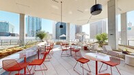 This dynamic rooftop amenity space offers a seamlessly integrated indoor-outdoor experience set against a dramatic urban skyline backdrop. The design is anchored by a bold, contemporary aesthetic — signature coral-orange wire-frame seating and tables are distributed throughout, creating a cohesive, energetic palette across every zone.

At the heart of the interior portion sits a striking central bar, clad in warm tiled finishes and encircled by matching bar stools. Above it, an elaborate suspended sculptural installation of orange rods and globe pendants soars toward the double-height ceiling, becoming an undeniable focal point. A classic green kamado grill nearby signals a serious dedication to outdoor entertaining.

The covered terrace transitions fluidly into open-air lounging areas where large concrete columns frame panoramic city views. Lounge chairs, modular sofas, and bistro-style seating arrangements cater to both relaxed socializing and casual dining. Native desert plantings and sculptural art pieces — including metallic figurines and a vivid violet wall medallion — add curated character throughout.

An upper terrace level extends the outdoor space further, furnished with additional seating under shade umbrellas with unobstructed skyline views on all sides. Overhead misters and fans ensure year-round comfort. The space is bathed in abundant natural light, making it as functional during the day as it is atmospheric at night.