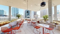 This dynamic rooftop amenity space offers a seamlessly integrated indoor-outdoor experience set against a dramatic urban skyline backdrop. The design is anchored by a bold, contemporary aesthetic — signature coral-orange wire-frame seating and tables are distributed throughout, creating a cohesive, energetic palette across every zone.

At the heart of the interior portion sits a striking central bar, clad in warm tiled finishes and encircled by matching bar stools. Above it, an elaborate suspended sculptural installation of orange rods and globe pendants soars toward the double-height ceiling, becoming an undeniable focal point. A classic green kamado grill nearby signals a serious dedication to outdoor entertaining.

The covered terrace transitions fluidly into open-air lounging areas where large concrete columns frame panoramic city views. Lounge chairs, modular sofas, and bistro-style seating arrangements cater to both relaxed socializing and casual dining. Native desert plantings and sculptural art pieces — including metallic figurines and a vivid violet wall medallion — add curated character throughout.

An upper terrace level extends the outdoor space further, furnished with additional seating under shade umbrellas with unobstructed skyline views on all sides. Overhead misters and fans ensure year-round comfort. The space is bathed in abundant natural light, making it as functional during the day as it is atmospheric at night.