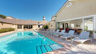 Located at 7099 North Hualapai Way in Las Vegas, NV, this single-story clubhouse serves as a comprehensive amenity hub designed to support an active and connected lifestyle. The outdoor areas are a highlight of the facility, showcasing two resort-style swimming pools surrounded by spacious pool decks furnished with lounge chairs and shaded umbrellas. A striking stone fireplace anchors one of the outdoor gathering areas, adding warmth and character to the desert setting beneath wide-open Nevada skies.

Inside, the fitness center is divided into two well-equipped zones: a cardio and strength machine room featuring treadmills, ellipticals, and cable machines, and a separate free weights and functional training area outfitted with a full dumbbell rack, TRX suspension system, and adjustable bench. Large windows and glass doors flood both areas with natural light and views of the surrounding community.

The clubhouse also includes a modern kitchen and dining space with dark cabinetry, tile backsplash, stainless steel appliances, and a standout live-edge wood dining table. A bright, open leasing or business office features floor-to-ceiling windows and transom glass, creating an airy and professional environment. Additional spaces include a media lounge with bold geometric tile flooring and a wall-mounted display screen, and a compact business center workstation for resident use.