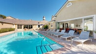 Located at 7099 North Hualapai Way in Las Vegas, NV, this single-story clubhouse serves as a comprehensive amenity hub designed to support an active and connected lifestyle. The outdoor areas are a highlight of the facility, showcasing two resort-style swimming pools surrounded by spacious pool decks furnished with lounge chairs and shaded umbrellas. A striking stone fireplace anchors one of the outdoor gathering areas, adding warmth and character to the desert setting beneath wide-open Nevada skies.

Inside, the fitness center is divided into two well-equipped zones: a cardio and strength machine room featuring treadmills, ellipticals, and cable machines, and a separate free weights and functional training area outfitted with a full dumbbell rack, TRX suspension system, and adjustable bench. Large windows and glass doors flood both areas with natural light and views of the surrounding community.

The clubhouse also includes a modern kitchen and dining space with dark cabinetry, tile backsplash, stainless steel appliances, and a standout live-edge wood dining table. A bright, open leasing or business office features floor-to-ceiling windows and transom glass, creating an airy and professional environment. Additional spaces include a media lounge with bold geometric tile flooring and a wall-mounted display screen, and a compact business center workstation for resident use.