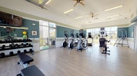 This fitness center is thoughtfully designed to support a comprehensive workout experience within a bright, welcoming environment. The space is characterized by a distinctive two-tone teal and white striped wall treatment, light wood-look flooring, and vaulted ceilings equipped with dual ceiling fans that keep air circulating throughout. Natural light floods the interior through large floor-to-ceiling windows on multiple walls, while an overhead garage-style door opens fully to a covered outdoor patio, seamlessly blending indoor and outdoor fitness.

The cardio zone features a row of Matrix elliptical trainers and treadmills, each equipped with individual monitors, positioned to face the expansive windows and enjoy views of the surrounding community. A dedicated strength and functional training area offers a rack of dumbbells in varying weights, kettlebells, a flat workout bench, stability balls, resistance bands, and a yoga/exercise guide board. A wall-mounted flat-screen TV serves this area as well.

An entry nook provides a small refreshment station with a mini-fridge, shelving, and decorative touches that create a welcoming arrival. The open garage door leads to a spacious covered outdoor patio with pergola-style roofing, overlooking a pool area and the surrounding residential buildings — ideal for outdoor stretching or cool-down sessions.