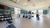 This fitness center is thoughtfully designed to support a comprehensive workout experience within a bright, welcoming environment. The space is characterized by a distinctive two-tone teal and white striped wall treatment, light wood-look flooring, and vaulted ceilings equipped with dual ceiling fans that keep air circulating throughout. Natural light floods the interior through large floor-to-ceiling windows on multiple walls, while an overhead garage-style door opens fully to a covered outdoor patio, seamlessly blending indoor and outdoor fitness.

The cardio zone features a row of Matrix elliptical trainers and treadmills, each equipped with individual monitors, positioned to face the expansive windows and enjoy views of the surrounding community. A dedicated strength and functional training area offers a rack of dumbbells in varying weights, kettlebells, a flat workout bench, stability balls, resistance bands, and a yoga/exercise guide board. A wall-mounted flat-screen TV serves this area as well.

An entry nook provides a small refreshment station with a mini-fridge, shelving, and decorative touches that create a welcoming arrival. The open garage door leads to a spacious covered outdoor patio with pergola-style roofing, overlooking a pool area and the surrounding residential buildings — ideal for outdoor stretching or cool-down sessions.