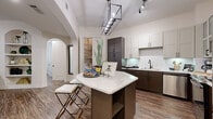 This single-story residence combines contemporary design with functional living across several well-defined spaces. The kitchen serves as a central highlight, featuring a two-tone cabinetry scheme pairing white upper cabinets with dark espresso lower cabinets, a classic white subway tile backsplash, and a curved quartz-topped island with integrated seating. Stainless steel appliances, a deep undermount sink with a matte black faucet, and a bold geometric pendant light complete the space. An arched doorway leads to a walk-in pantry, adding both character and practicality.

The living area showcases vaulted ceilings with crown molding, wood-look plank flooring, and a wall of large windows with access to a balcony, flooding the room with natural light. Built-in shelving flanks a media console, providing ample display and storage space. A ceiling fan enhances the airy feel.

Both bedrooms are comfortably sized, each featuring wood-look flooring, natural light from windowed walls with wood-framed blinds, and ample wall space. The bathroom features a full soaking tub with white subway tile surround, tile flooring, and a bold black-and-white striped shower curtain that echoes the home's consistent design palette.