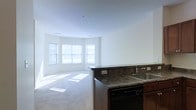 This single-floor space showcases an open-concept layout that integrates the kitchen and living area into one cohesive environment. The kitchen is equipped with rich, dark wood cabinetry and deep granite countertops that extend along a peninsula, which serves as a natural divider between the cooking and living zones. A stainless steel sink is positioned along the countertop, and a built-in black dishwasher is neatly integrated beneath the cabinetry. Electrical outlets are conveniently placed along the peninsula's side, adding functional utility to the space. The living area features plush wall-to-wall carpeting that contributes to a warm, comfortable atmosphere. A curved bay window arrangement along the far wall allows generous amounts of natural light to flood the interior, casting bright patterns across the floor. The walls are finished in a clean, neutral white tone that enhances the sense of openness throughout. Smooth, flat ceilings complement the minimalist aesthetic, and the overall layout offers a practical, airy feel well-suited for everyday living.
