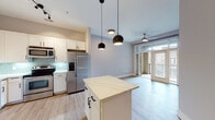Located at 600 St. Mary's St in Raleigh, NC, this well-appointed single-floor residence offers a thoughtfully designed open-concept layout that seamlessly connects the kitchen, dining, and living areas. The kitchen is a standout feature, outfitted with stainless steel appliances including a French door refrigerator, electric range, microwave, and dishwasher. Crisp white shaker-style cabinetry, a soft aqua glass tile backsplash, and light hardwood flooring lend the space a clean, contemporary aesthetic. A central island with a white quartz countertop provides additional prep space and serves as a natural gathering point. Stylish spherical pendant lights hang above the island, complementing the ceiling fan in the adjacent living area, which opens to a private balcony through large glass doors, allowing ample natural light to flow through. The bedroom features plush carpeting, a ceiling fan, and a window with natural light, while an en-suite bathroom with dark wood cabinetry and tile flooring adds a refined touch. A dedicated in-unit laundry closet with a stacked washer and dryer adds everyday convenience. Soft gray walls throughout create a calm, neutral palette that ties the entire space together cohesively.