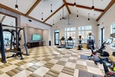 This modern fitness center offers a spacious, single-floor layout designed to support a comprehensive workout experience. The architectural centerpiece is the dramatic vaulted ceiling adorned with exposed wood beams, which lend a warm, lodge-inspired character to the space. Cylindrical pendant lights hang at varying heights, complementing the ceiling fans that keep the environment comfortable during workouts.

The main exercise area features a patterned carpet tile floor in a checkered arrangement of neutral tones, providing both durability and visual interest. Cardio equipment, including treadmills, stationary bikes, a stair climber, and cycling bikes, lines the perimeter, while a multi-station cable machine and free weights cater to strength training needs. A large wall-mounted flat-screen television is set against a distinctive wood-panel accent wall, with open storage cubbies beneath — ideal for personal items.

Floor-to-ceiling glass doors and a generous bank of windows along multiple walls flood the space with natural light and offer views of the surrounding wooded landscape. A full-length mirror wall amplifies the sense of openness. A connecting hallway — adorned with framed artwork — leads into the fitness area, adding a refined, gallery-like entry experience to the overall welcoming and motivating atmosphere.