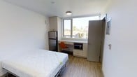 This efficiently designed studio unit offers a thoughtful layout that maximizes every inch of its compact footprint. The main living area serves multiple functions, combining sleeping, working, and living spaces into a single, well-organized room. Light hardwood-style flooring runs throughout, contributing to a clean and modern aesthetic.

A large window positioned along the exterior wall floods the space with natural light and frames views of the surrounding neighborhood rooftops and greenery. Just beneath the window sits a built-in desk with lower cabinet storage, providing a dedicated workspace. Adjacent to the desk, a full-size stainless steel refrigerator is neatly integrated alongside upper cabinetry that offers additional storage. A microwave is conveniently housed within the lower cabinetry unit.

The room features white walls and a smooth ceiling with a centrally mounted light fixture, keeping the overall ambiance bright and neutral. A sliding door leads to a closet area on one side of the room, while another door provides access to the private bathroom. The space is designed with functionality and simplicity in mind, making it a practical and self-contained living environment suited for individual occupancy.