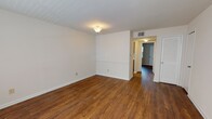 Located at 115 Stratford Rd in Williamsburg, Virginia, this single-floor unit offers a straightforward, functional layout with consistent design elements throughout. Rich, warm-toned hardwood-style flooring runs across the living areas and bedrooms, providing a cohesive aesthetic that ties the space together.

The living area is generously proportioned, featuring white walls with subtle horizontal detailing and a louvered closet door that adds a classic touch. A hallway connects the main living space to the rest of the unit, with overhead lighting illuminating the path. The layout, as seen in the floor plan, flows efficiently from the living zone through to the sleeping areas and kitchen.

The bedrooms are well-sized, each featuring windows fitted with horizontal blinds that allow natural light to filter in while maintaining privacy. The neutral wall tones and continued hardwood-style flooring keep the rooms feeling bright and consistent.

The kitchen is equipped with warm oak cabinetry, white countertops, a full-size refrigerator, and an electric range with an overhead ventilation hood, providing essential functionality in a compact footprint.

The bathroom features a tiled tub-shower surround in a neutral beige palette, a white vanity with countertop sink, and hardwood-style flooring, creating a clean, practical space.