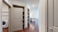 This single-floor residence showcases a thoughtfully designed, open-concept layout with a seamless flow between the main living areas. Rich, dark hardwood flooring runs throughout, providing a warm contrast to the crisp white walls and trim.

The kitchen serves as a central hub, featuring shaker-style cabinetry in a soft gray-white finish, a white subway tile backsplash, stainless steel appliances including a full-size refrigerator and range, and a spacious central island with a light quartz countertop. Pendant lighting adds a refined touch above the island and dining area. An open built-in shelving unit adjacent to the kitchen offers additional storage and display space, along with a dedicated pantry closet nearby.

The living area is generously proportioned, illuminated by natural light from double doors and complemented by a ceiling fan. A hallway leads to the bathroom, which features a wide vanity with an undermount sink, a large mirror, dark navy subway tile surrounding the soaking tub, and a frosted privacy window. The dedicated laundry area accommodates a full-size side-by-side washer and dryer beneath wall-mounted shelving, adding practical convenience to the space.