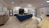 This single-floor space is designed as a contemporary communal lounge and dining area, combining functionality with modern aesthetic sensibility. The open-plan layout creates a generous, airy feel, with light wood-style flooring running throughout and contributing to a warm, cohesive atmosphere.

The lounge area is anchored by a large navy blue sectional sofa, positioned atop a gold-and-grey abstract area rug. Opposite the seating, a dramatic floor-to-ceiling entertainment wall in matte black houses a large flat-screen television, integrated shelving, and ample cabinetry storage. Track lighting overhead provides adjustable, gallery-style illumination throughout the space.

A dedicated dining area sits adjacent to the lounge, centered around a substantial wood table surrounded by upholstered chairs. A large decorative mirror with an ornate frame is mounted on the wall above, enhancing the sense of depth and light. Natural light filters in through multiple windows, offering views of the surrounding streetscape.

A quieter seating nook near the windows features a pair of charcoal upholstered accent chairs alongside textured wall art, offering a more intimate setting within the broader open space.