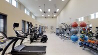 This fitness center offers a comprehensive and thoughtfully designed workout environment spread across a single level. The space is defined by its impressively high ceilings, which lend an open, airy feel throughout. Natural light streams in through a row of clerestory windows positioned along the upper walls, while recessed and pendant-style lighting ensures the interior remains bright and welcoming at all hours.

The main exercise floor is generously sized and features a polished, easy-to-maintain surface. A full lineup of SportsArt cardio machines — including treadmills and elliptical trainers — is arranged in orderly rows, offering ample space between each unit for comfortable use. Along one side, a mirrored wall visually expands the room and supports form-checking during workouts. Stability balls, free weights, and a dedicated strength training area round out the equipment offerings, catering to a wide variety of fitness routines.

Framed artwork along the walls adds a subtle aesthetic touch, while ceiling fans help maintain comfortable air circulation throughout the space. The overall ambiance is clean, functional, and motivating — well-suited for both casual exercisers and dedicated fitness enthusiasts alike.