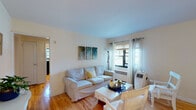 This single-floor residence at Bayshore Manor in Bay Shore, NY offers a well-organized layout with distinct living, dining, sleeping, and cooking areas connected by warm hardwood flooring. The living room is a comfortable, light-filled space featuring large windows dressed with sheer curtains that allow natural light to filter in. A neutral-toned sofa, glass-top coffee table, and wall-mounted artwork contribute to the room's relaxed, cohesive aesthetic. The living area flows directly into a dining space furnished with a wooden table and chairs, alongside a flat-screen television setup.

The kitchen is a compact, functional galley-style space equipped with wooden cabinetry, dark countertops, a stainless steel refrigerator, and a window that provides natural light and outdoor views. Dark vinyl flooring distinguishes it from the rest of the unit.

Two bedrooms provide varied sleeping configurations. The primary bedroom features hardwood floors, multiple windows with draping curtains, a full dresser, and wall-mounted television. The second bedroom accommodates bunk beds alongside a twin bed, making it a versatile space. The bathroom is clean and simply appointed with a white vanity, mirrored medicine cabinet, and a shower-tub combination.