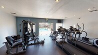 Located in Fountain Hills, Arizona, this single-floor fitness center offers a well-equipped workout facility designed to support a range of exercise routines. The space is finished with durable rubber flooring throughout, providing a practical and comfortable surface for training. Walls are painted in a clean combination of soft blue and white tones, lending the room a fresh, energizing atmosphere complemented by recessed lighting and a textured ceiling.

The cardio area features multiple treadmills and an elliptical machine, positioned to take advantage of natural light and views of the outdoor pool area beyond a set of large glass French doors. This direct visual connection to the pool creates a bright, open feel within the fitness space. A wall-mounted mirror near the free weight station enhances the sense of depth, while a dumbbell rack provides a organized storage solution for hand weights.

Strength training is well represented by a multi-station BATCA cable machine, offering a variety of resistance-based exercises within a compact footprint. An additional dedicated area for strength equipment provides further versatility for users. The entry corridor, finished in light blue with durable flooring, leads directly into the main workout area, with a window looking into an adjacent office or amenity space. The fitness center is conveniently accessible and thoughtfully laid out for community use.