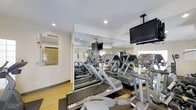 This fitness center offers a comprehensive and well-organized workout environment designed to meet a variety of exercise needs. The space is outfitted with a selection of cardio equipment, including treadmills and elliptical trainers, arranged to allow comfortable use and easy movement between machines. Large wall-to-wall mirrors line one side of the room, creating a sense of expanded space while allowing users to monitor their form during exercise. Ceiling-mounted flat-screen televisions are positioned for easy viewing from the cardio equipment, adding an entertainment element to workouts. The flooring is a light-toned hardwood-style surface that contributes to the clean, polished aesthetic of the room. Recessed lighting in the white ceiling provides bright, even illumination throughout. A wall-mounted air conditioning unit ensures a comfortable climate during use. An informational and emergency procedures notice is prominently displayed on the wall, reflecting attention to safety. Natural light filters in through windows, further enhancing the open and energizing atmosphere. Overall, this fitness center presents a functional, well-maintained space suited for daily cardiovascular and general fitness activities.