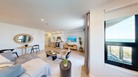 Located at The Breeze on East Broadway in Long Beach, NY, this thoughtfully designed unit offers a modern coastal aesthetic throughout its single-floor layout. Light wood-tone flooring flows seamlessly across the open-concept living and kitchen area, creating a bright and cohesive atmosphere enhanced by recessed lighting and crisp white walls.

The living area is generously proportioned, featuring large windows and a private balcony that frames views of the surrounding cityscape and glimpses of the water beyond. Sheer drapery softens the natural light that fills the space throughout the day.

The kitchen is efficiently designed with white cabinetry accented by matte black hardware, stainless steel appliances including a refrigerator, range, and microwave, and a clean white countertop. Upper cabinetry extends to the ceiling for ample storage. A compact dining area alongside the kitchen adds functional versatility to the space.

The bathroom is finished to a high standard, featuring a wood-toned vanity with a white countertop, an illuminated LED mirror, and a glass-enclosed walk-in shower with white subway tile and a rain showerhead. Neutral gray wall tones complement the warm wood accents.

The hallway provides a well-organized entry with additional upper cabinetry, offering practical storage from the moment you step inside.