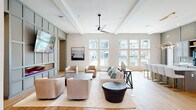 Located along South Blue Angel Parkway in Pensacola, Florida, this single-story clubhouse is designed as a contemporary community amenity space with a cohesive modern aesthetic throughout. Light hardwood flooring, neutral tones, and carefully selected furnishings create a polished yet approachable atmosphere across all areas.

The main lounge is a generously proportioned gathering space anchored by a coffered ceiling with a sleek ceiling fan and an expansive bank of tall windows overlooking the outdoor pool area, flooding the room with natural light. Comfortable seating arrangements, including plush sofas and swivel chairs, are organized around area rugs, encouraging relaxed socializing. A mounted television and built-in cabinetry accent one wall.

Adjacent to the lounge is a well-appointed kitchen and coffee bar area featuring gray cabinetry, white quartz countertops, a central island with upholstered gold-accented bar stools, and a stylish layered pendant light fixture overhead.

A dedicated conference and business center offers a round table with ergonomic seating, a workstation, and bold aviation-themed artwork beneath dramatic drum pendant lighting.

The fitness center is equipped with a full range of strength and cardio machines, rubber flooring, mirrored walls, and large windows providing natural light and views of the surrounding landscape.

A covered outdoor terrace connects seamlessly to the pool deck, furnished with modern lounge seating and large ceiling fans.
